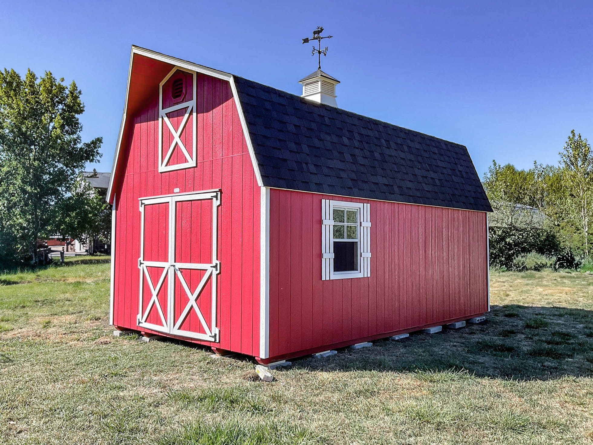Bright red barn-style shed with white trim, featuring double doors and a steep gambrel roof, situated on a grassy field under a clear sky.