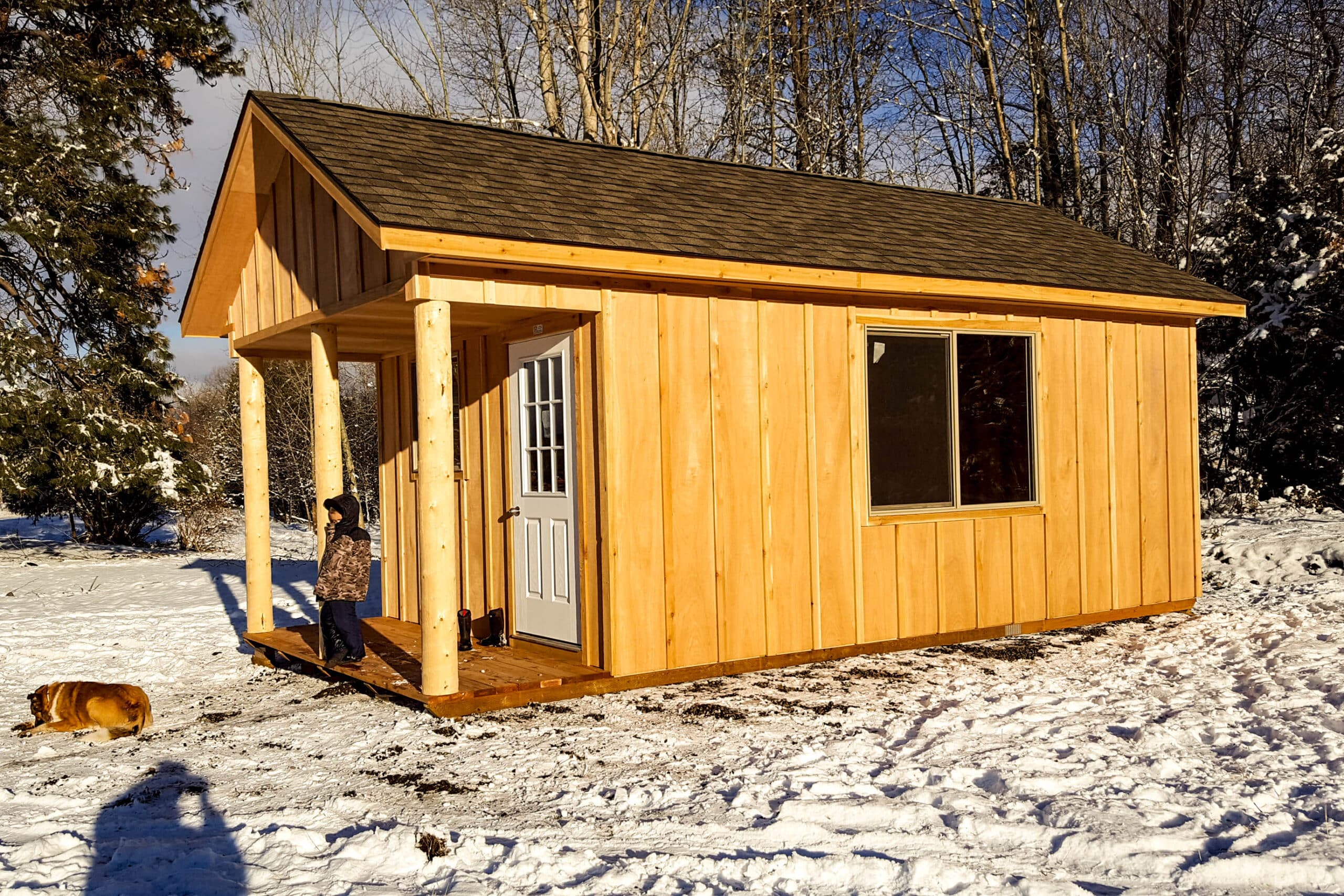 A wooden cabin with a front porch, set on a snowy ground surrounded by bare trees under a sunny sky.