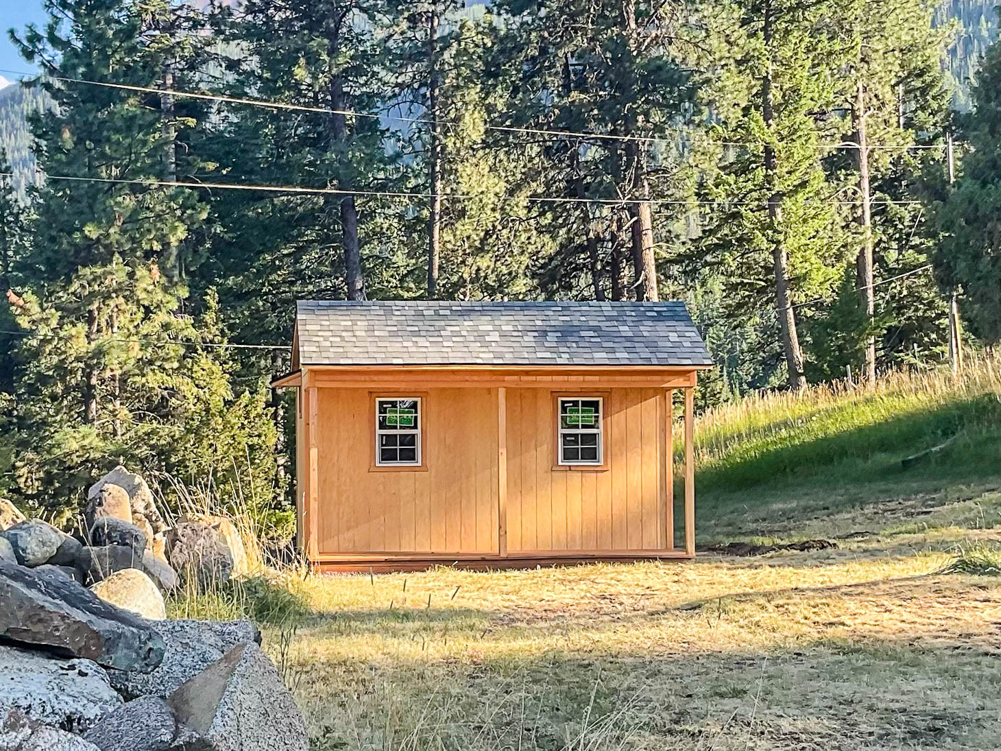 A compact wooden shed with a sloped shingled roof, a light natural wood finish, and two small windows. The shed sits on a gentle grassy slope, surrounded by tall trees and soft sunlight.