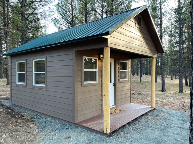 A brown cabin shed with a green metal gable roof, a small covered porch with wooden posts, and multiple windows with white trim. The shed is situated on a well-maintained grassy lot with a backdrop of pine trees and open skies. Just one of our popular cabins for sale in Pendleton.