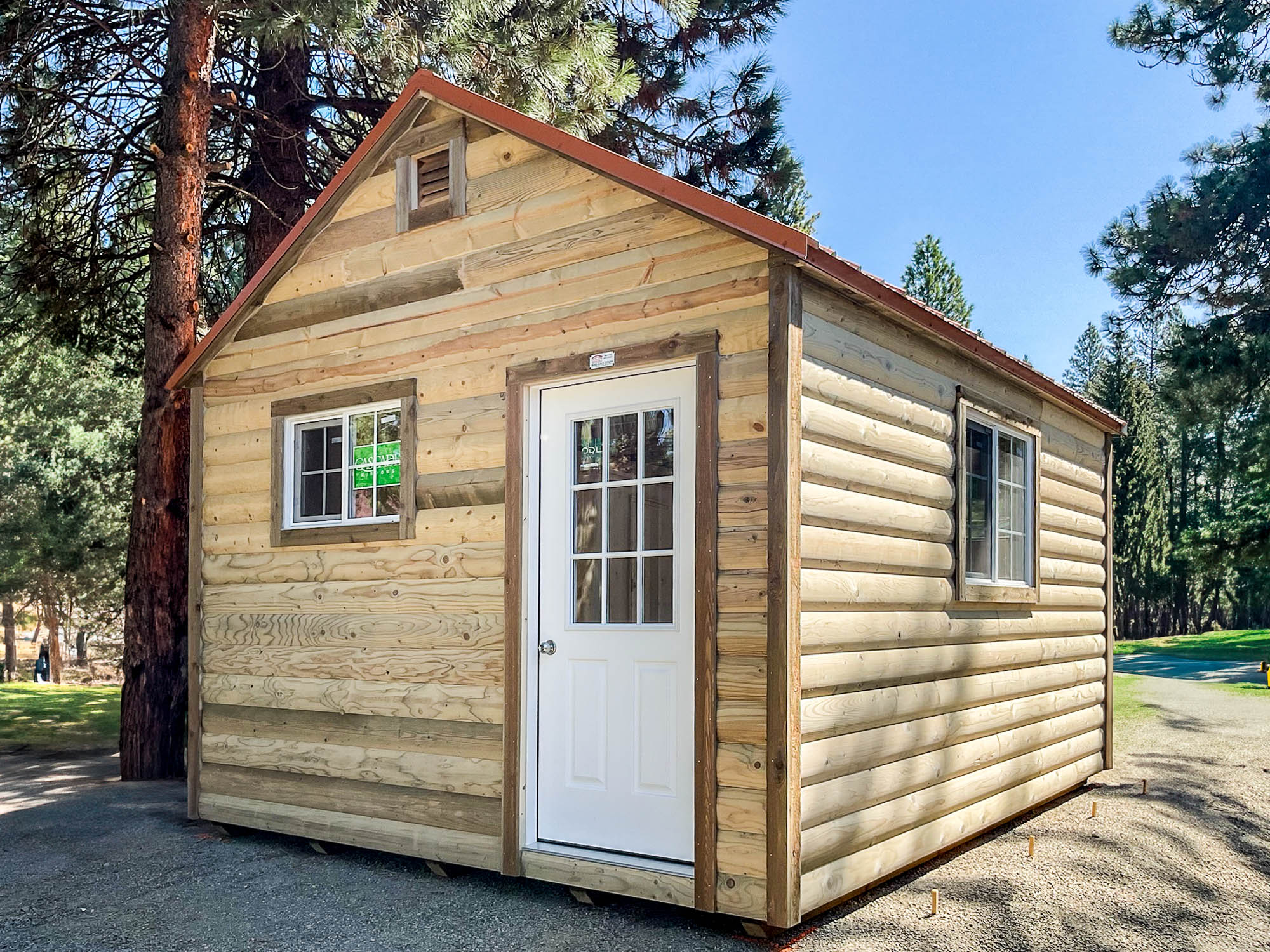 A tall, light-colored wooden gable shed with a vertical siding design, a shingled roof, a white entry door with a small window, and an additional side window. The shed is placed on gravel, surrounded by trees and open sky.