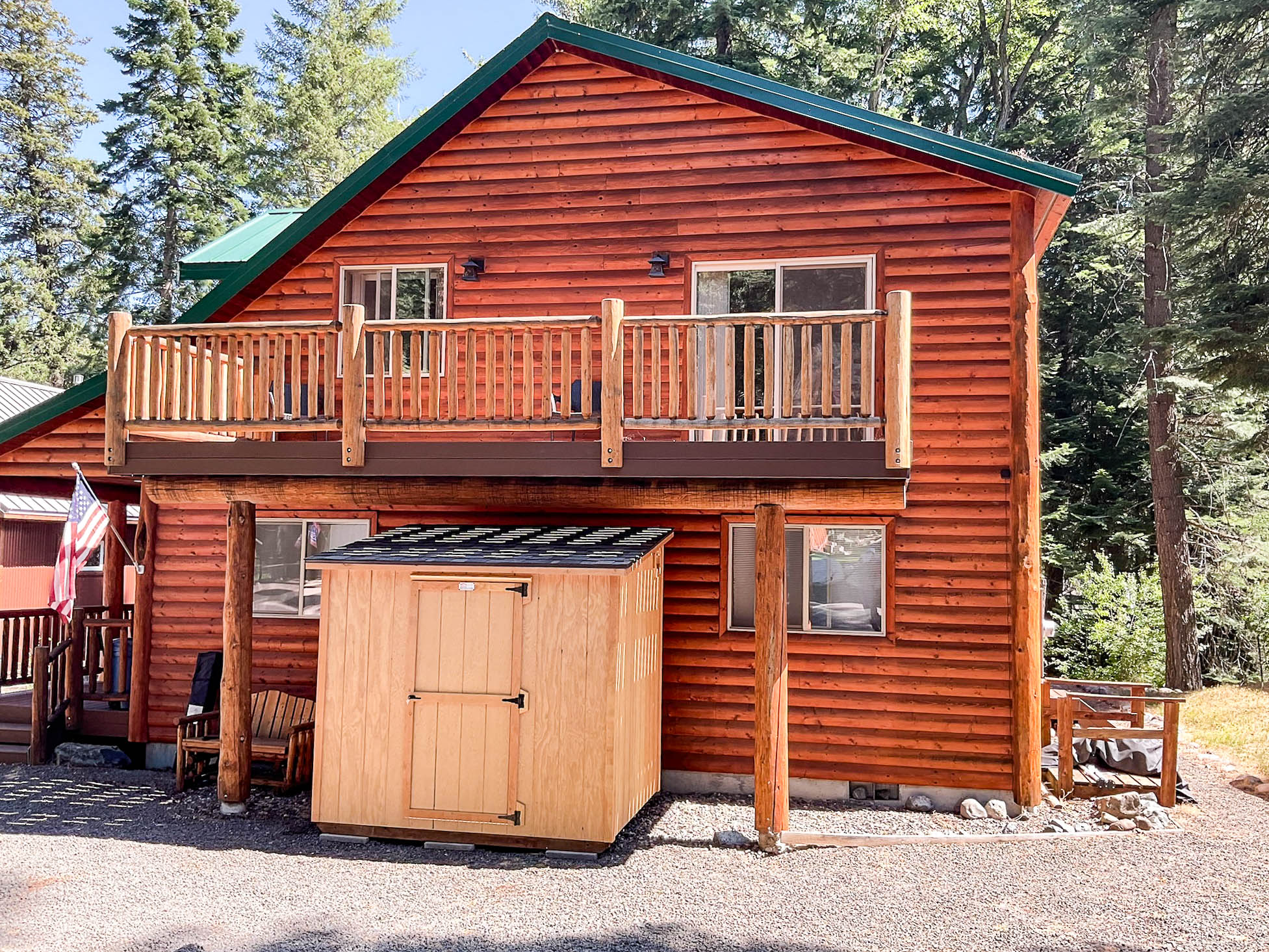 A wooden shed positioned under a second-story balcony of a log-style house with red siding and green trim.