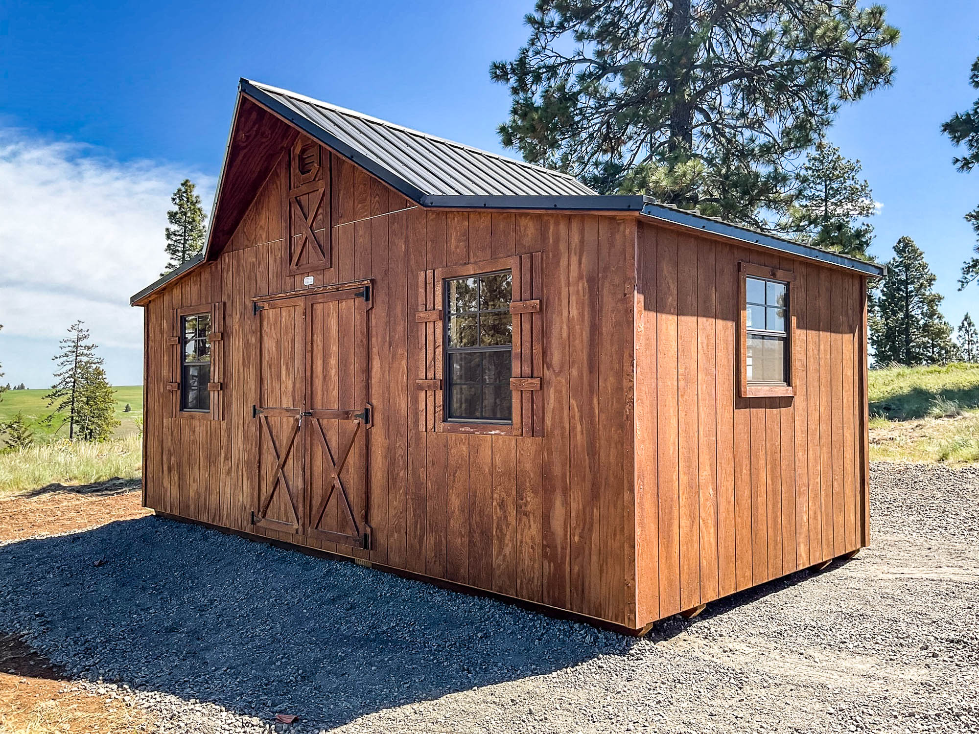 A wooden cabin-style shed with a natural cedar finish, a dark shingled roof, and multiple windows with brown trim. The shed has a single entry door and is placed on a gravel surface with a tall tree in the background. The warm-toned wood siding gives it a rustic appearance. One of our Western Barn sheds for sale in Pendleton.