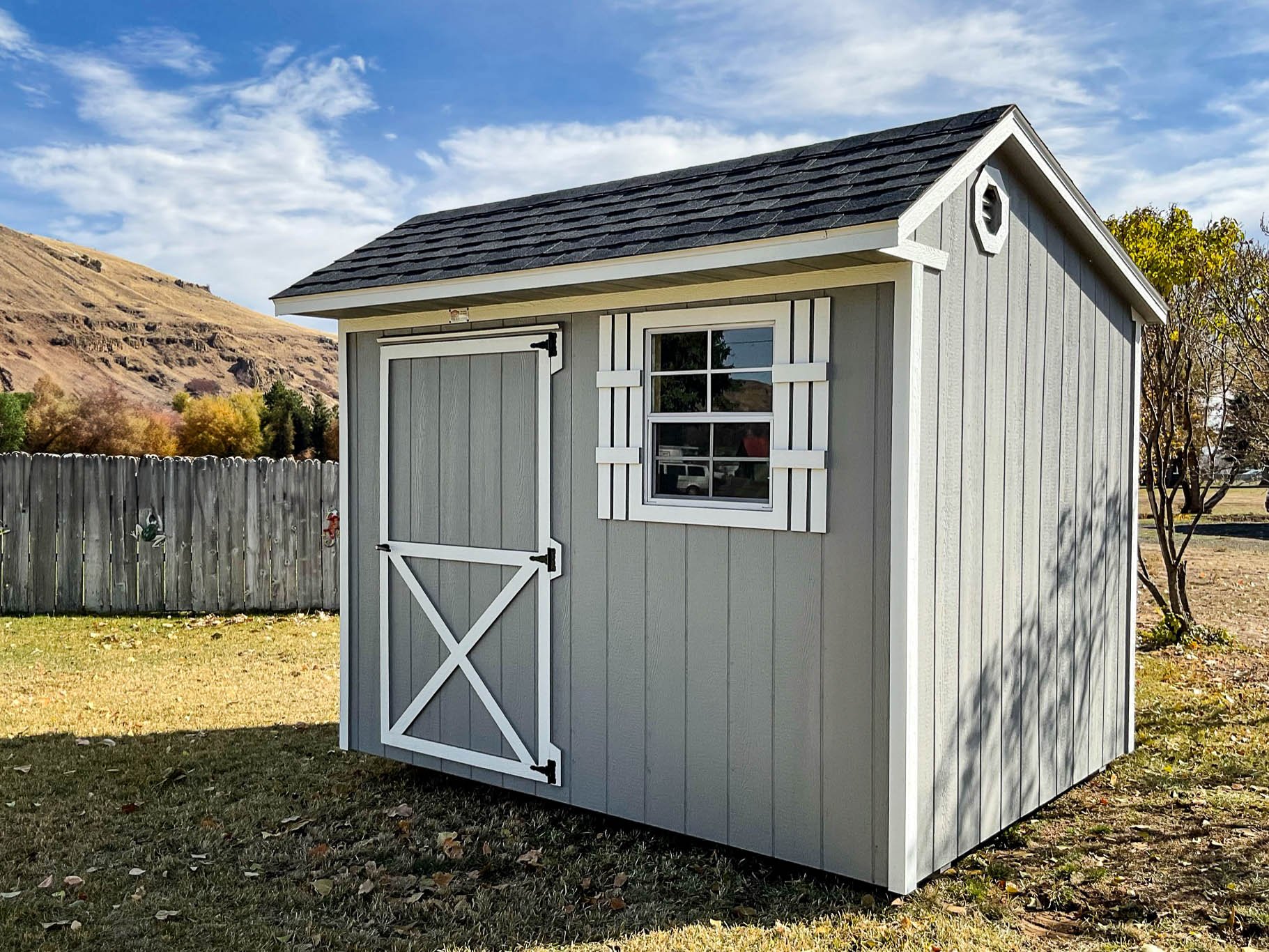 A light gray gable-style shed with a dark gray shingled roof, white trim, and a white barn-style single door featuring a cross-brace design. A small window with white trim is located on the side. The shed is positioned on a well-maintained grassy area with a wooden fence and rolling hills in the background. One of our compact sheds for sale in Pendleton.