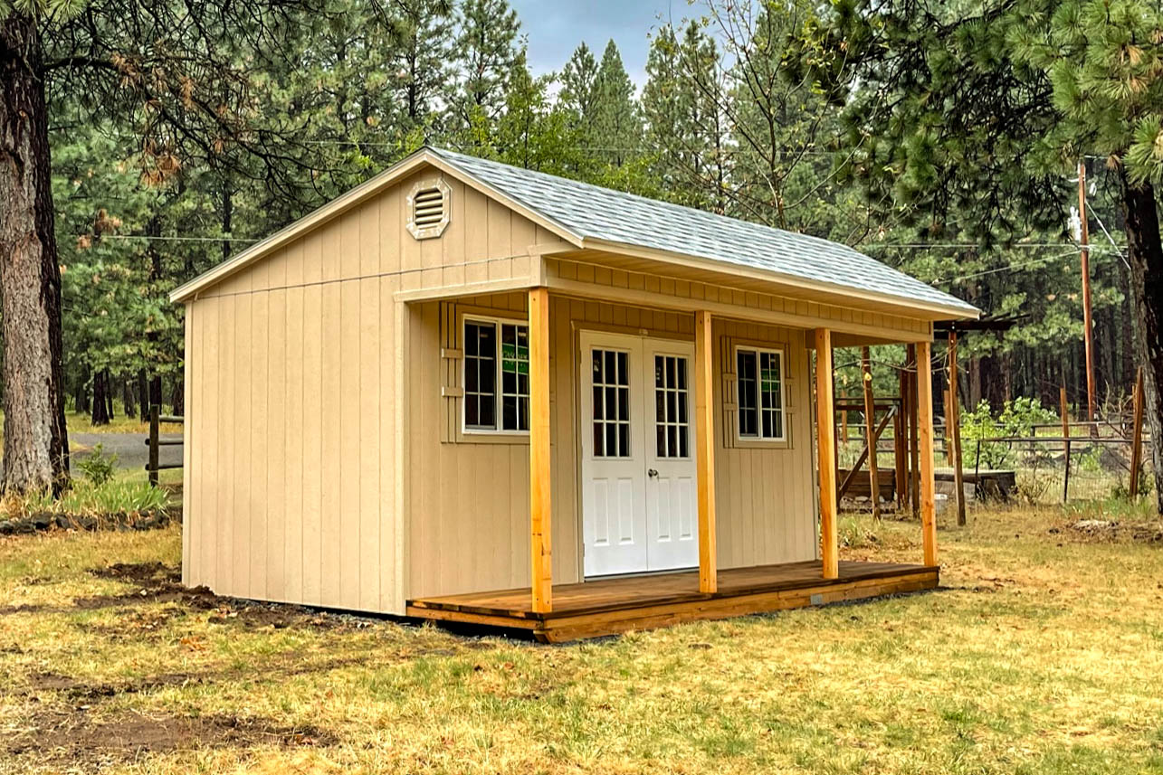 A beige cabin with a gray shingled roof, a front porch supported by wooden posts, and a white double door, set in a forested area.