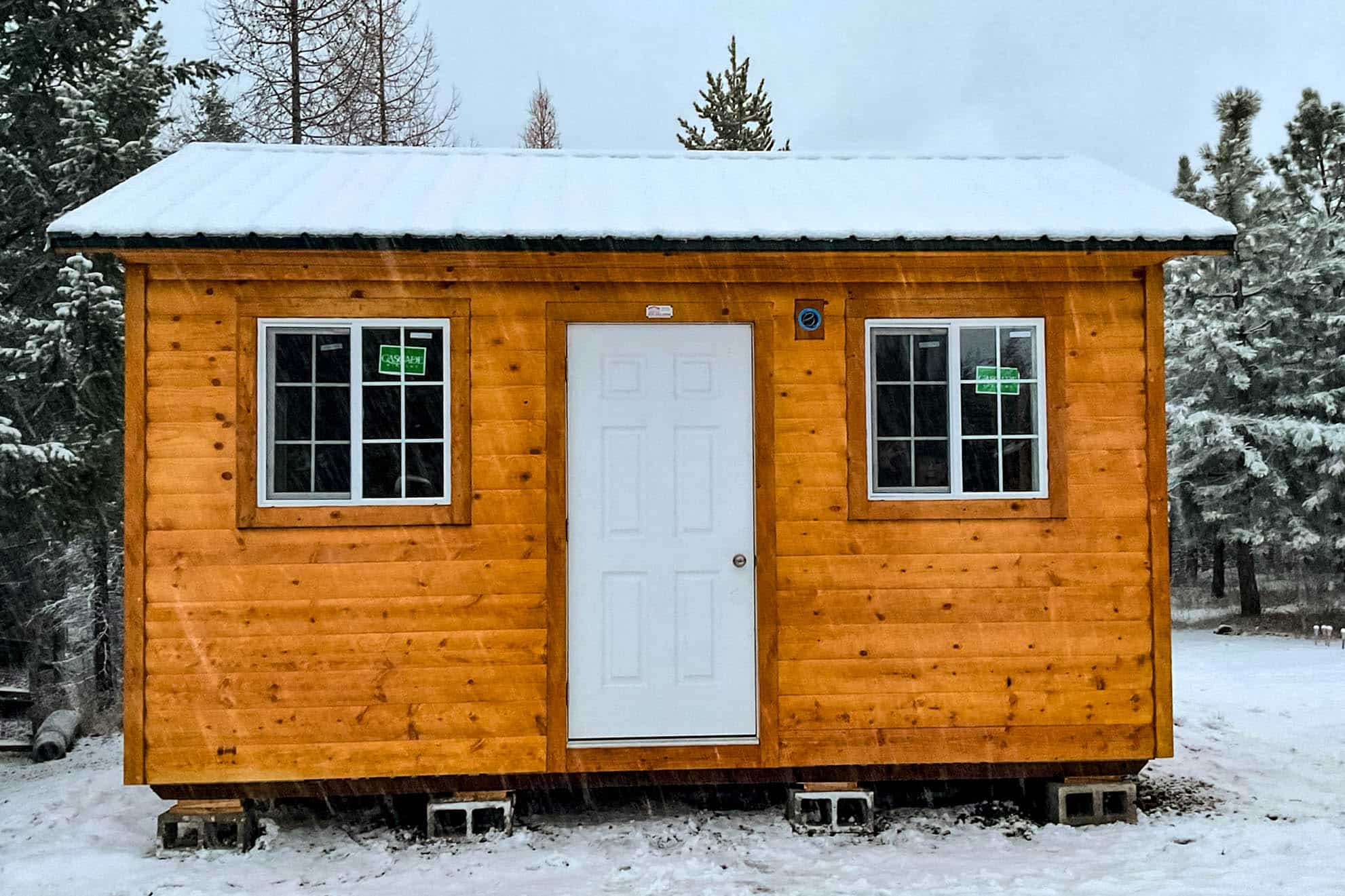 A small orange-toned wooden cabin with a white door and two windows, set in a snowy landscape surrounded by evergreen trees.