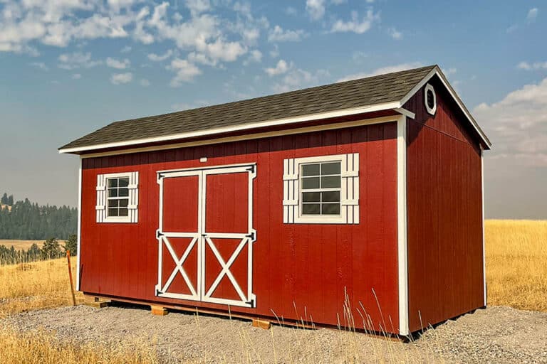 One of our Storage sheds for sale in Ontario - A red utility shed with white trim, barn-style double doors, and two side windows, set in a golden field under a partly cloudy sky.