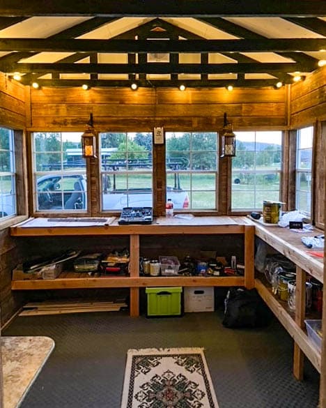 Interior of a rustic she shed with earthy wood tones, illuminated by warm string lights and lanterns, featuring workbenches, storage shelves, and a view of a green landscape through large windows.