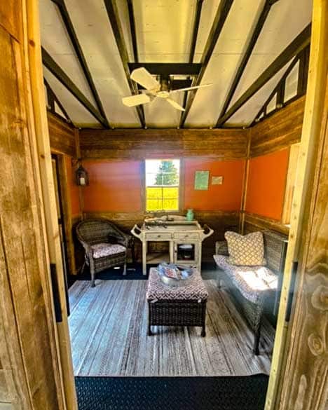 Cozy she shed interior with earthy burnt-orange walls, rustic wood accents, wicker furniture, and a white ceiling fan, illuminated by natural light streaming through a window.