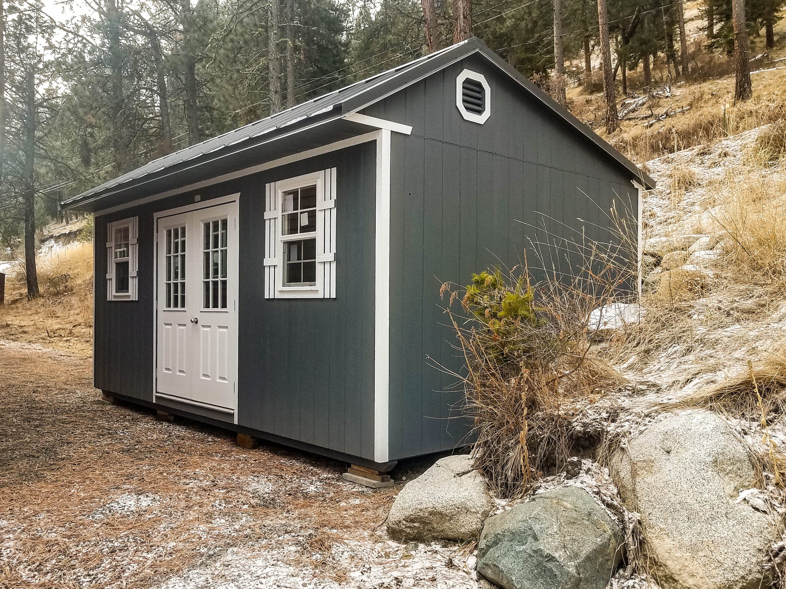 Dark gray tiny home shed with white trim, set in a natural outdoor setting surrounded by trees.