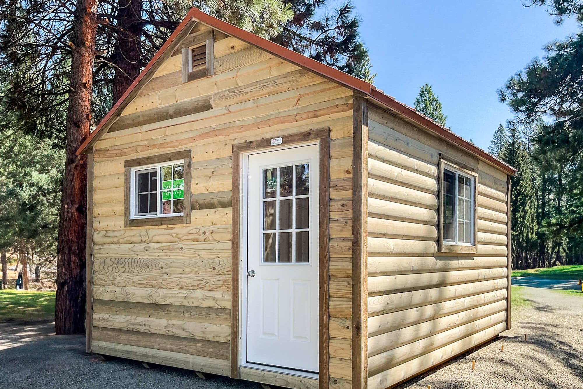 A tall, light-colored wooden gable shed with a vertical siding design, a shingled roof, a white entry door with a small window, and an additional side window. The shed is placed on gravel, surrounded by trees and open sky.