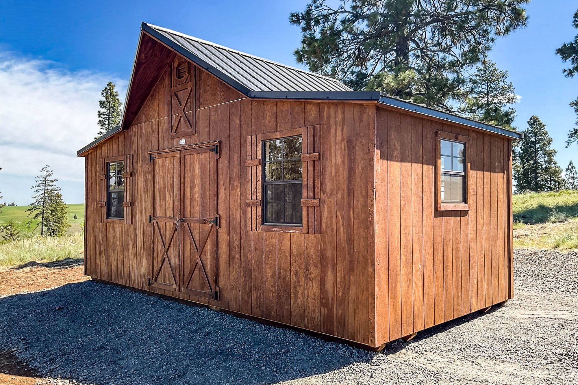 A wooden gable-style shed with a rich brown stained finish, a dark brown shingled roof, and two windows with brown trim. The shed is placed on a gravel foundation with a backdrop of tall evergreen trees and a clear blue sky.