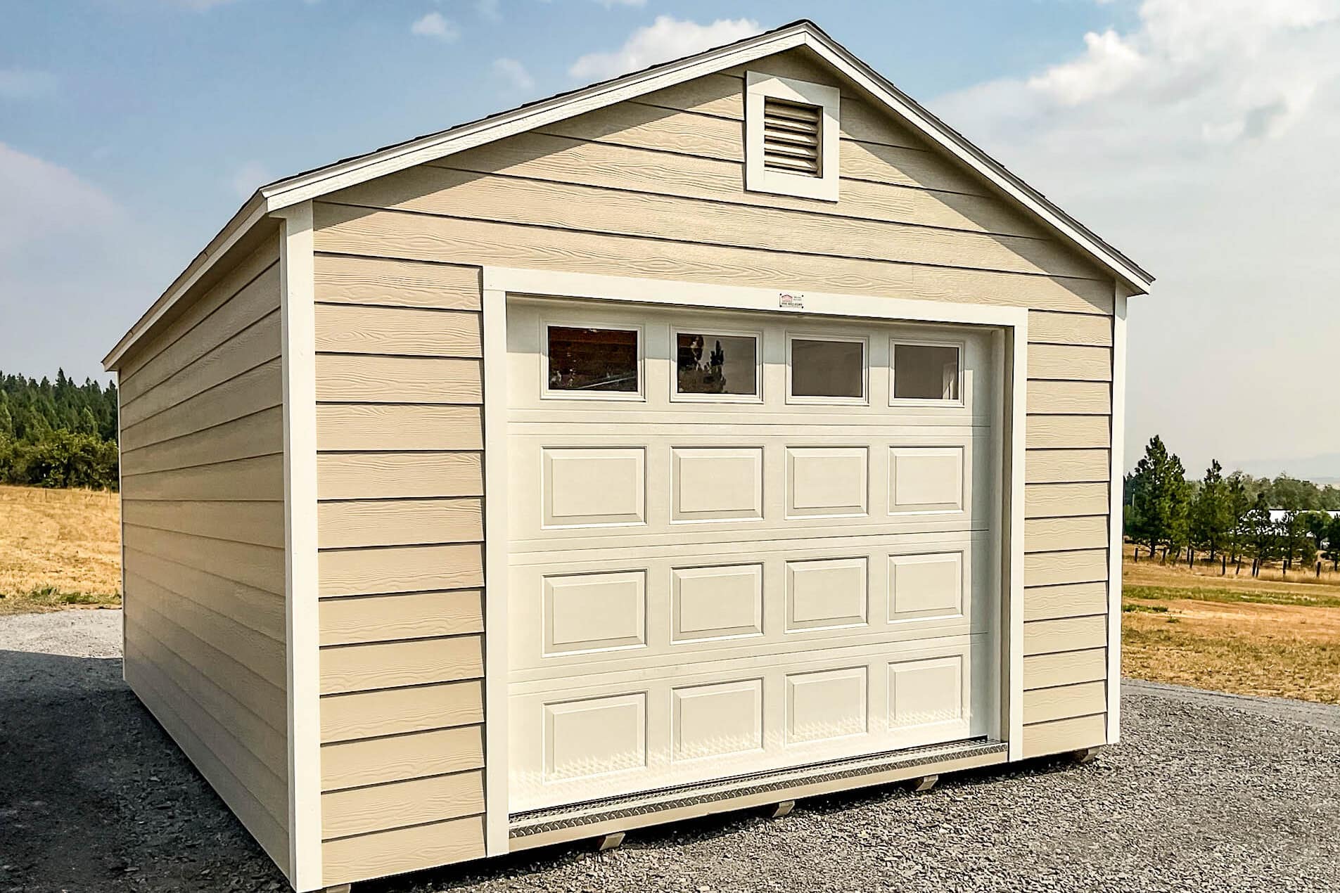 A beige gable-style shed with a dark shingled roof, white trim, and a large garage-style door with decorative windows. A small loft window sits at the peak of the gable, adding a functional and aesthetic touch. The shed is placed on a level gravel surface with an open landscape in the background. One of our garage sheds for sale in Pendleton.