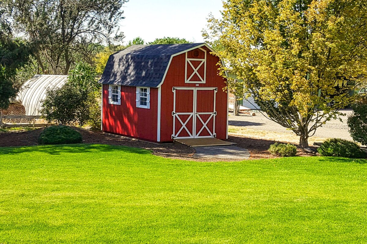 10x16 tall barn - Red painted wood siding with white-trimmed double doors with X detail on front - two windows with white shutters on left side - gambrel-style black shingled roof - barn is nestled among trees and faces neat green field