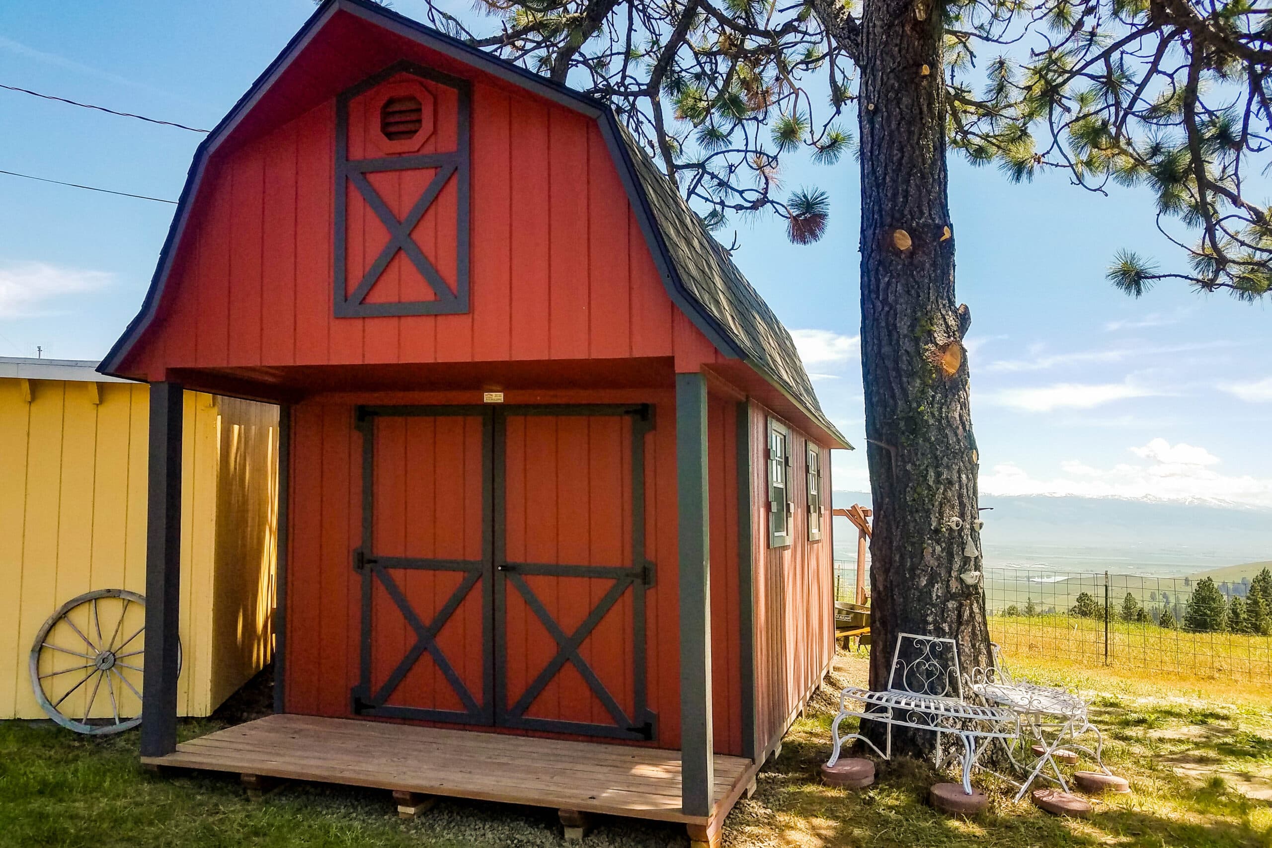 A red barn-style shed with dark gray trim, large double doors, and a loft area, set near a tree with a scenic outdoor backdrop